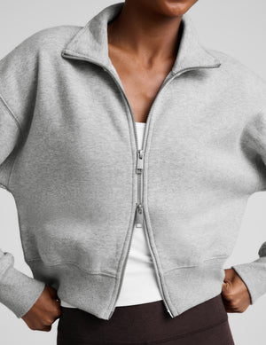A woman modeling a light gray zip-up fleece jacket and dark brown capri leggings against a light gray studio background.