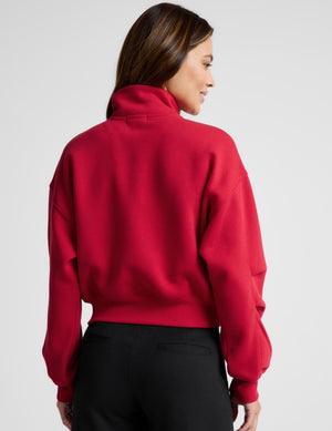 Woman wearing a red zip-up sweatshirt and black pants against a plain background. 