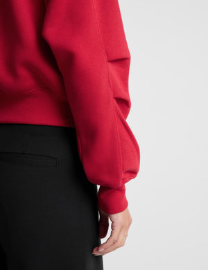 Woman wearing a red zip-up sweatshirt and black pants against a plain background. 