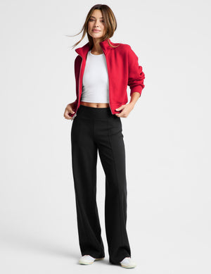 Woman wearing a red zip-up sweatshirt and black pants against a plain background. 