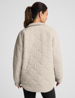 A woman shown from the back wearing a light beige quilted jacket with a wavy stitched pattern and ribbed collar, cuffs, and hem, against a plain light gray studio background.