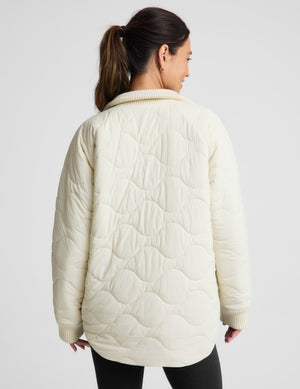 


A woman shown from the back wearing a cream-colored quilted jacket with a wavy stitched pattern and ribbed collar, cuffs, and hem, paired with black pants against a light gray studio background.