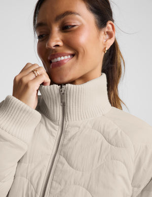 A close-up of a woman wearing a light beige quilted puffer jacket with a ribbed mock neck and front zipper, set against a plain light gray background.