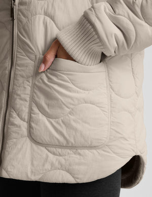 Close-up of a quilted beige puffer jacket with a large front patch pocket and a ribbed knit sleeve cuff, shown on a model with a hand in the pocket.