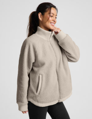 A woman wearing a light beige sherpa fleece zip-up jacket with a mock neck and ribbed cuffs, paired with black leggings, photographed against a solid light gray background.