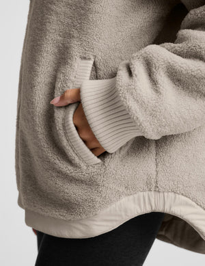 Close-up of a person wearing a soft taupe fleece jacket with a ribbed knit cuff and a side pocket, with a hand reaching into the pocket.