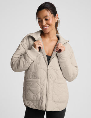 A woman wearing a light beige quilted zip-up jacket with a stand-up collar and front pockets, photographed against a plain light gray studio background.