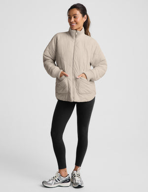 A woman modeling a light beige quilted zip-up jacket with pockets, paired with black leggings and silver sneakers, against a plain light gray studio background.