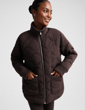 A woman wearing a dark brown quilted puffer jacket with a stand-up collar, front zipper, and ribbed cuffs, standing against a light gray studio background.