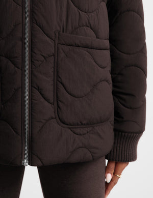 Close-up of a dark brown quilted puffer jacket with a wavy stitched pattern, ribbed cuffs, and a front zipper, worn by a model.