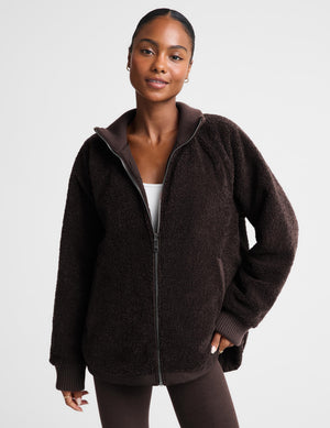 A woman modeling a dark brown sherpa zip-up hoodie jacket against a light gray studio background.