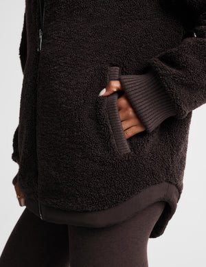 Close-up of a person wearing a dark brown sherpa fleece zip-up jacket with ribbed knit cuffs and a matching dark brown ribbed hem, paired with dark leggings against a light gray background.