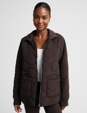 A woman wearing a dark brown quilted zip-up jacket with a ribbed collar, cuffs, and hem, layered over a white top and dark pants, photographed against a light gray studio background.