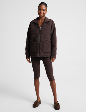 A woman modeling a dark brown quilted zip-up jacket, matching brown cropped leggings, and brown suede loafers against a light gray studio background.