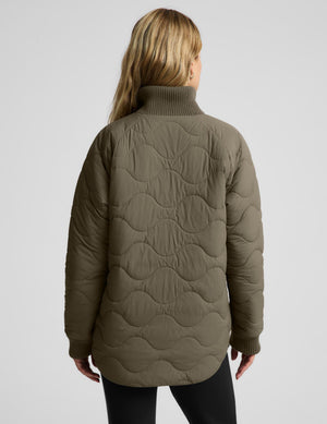 A woman viewed from the back wearing a quilted olive green turtleneck jacket against a light gray studio background.