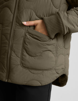 Close-up of a quilted olive green jacket with a hand in a pocket, showing the texture and ribbed sleeve cuff against a light gray background.