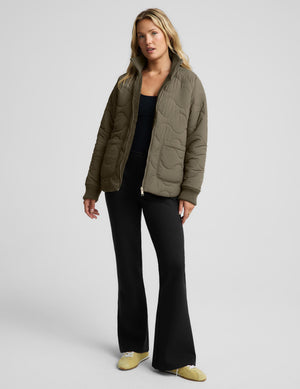 A woman models a quilted olive green jacket over a black top and black flared pants, paired with light yellow shoes, against a solid light gray studio background.