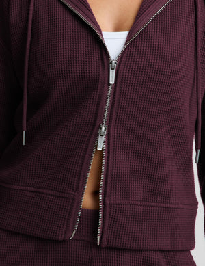 A woman modeling a matching burgundy waffle-knit zip-up hoodie and sweatpants set against a light gray studio background.