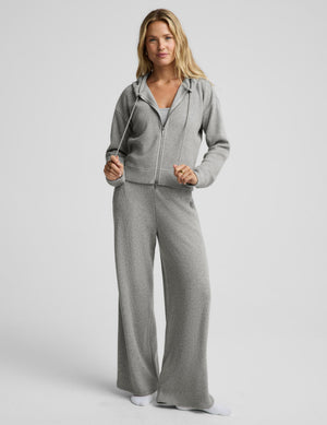 A woman modeling a light gray two-piece loungewear set consisting of a zip-up hoodie and matching wide-leg sweatpants, photographed against a solid light gray studio background.