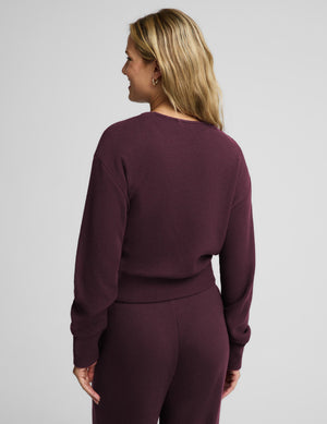 A smiling blonde woman wearing a matching deep plum purple ribbed knit loungewear set, featuring a wrap-style cardigan with a tie belt and coordinating pants, against a solid light gray background.