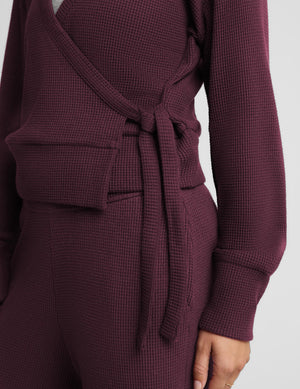 A blonde woman wearing a matching deep plum purple ribbed knit loungewear set, featuring a wrap-style cardigan with a tie belt and coordinating pants, against a solid light gray background.