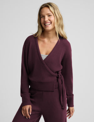 A smiling blonde woman wearing a matching deep plum purple ribbed knit loungewear set, featuring a wrap-style cardigan with a tie belt and coordinating pants, against a solid light gray background.