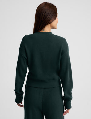 A woman wearing a dark forest green knit wrap cardigan with a tie waist and matching knit pants, photographed against a light gray studio background.