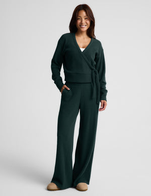 A woman wearing a dark forest green knit wrap cardigan with a tie waist and matching knit pants, photographed against a light gray studio background.