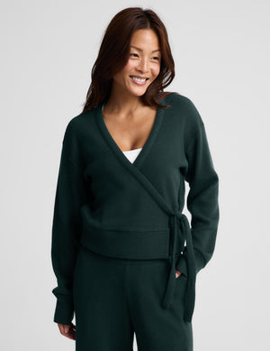 A woman wearing a dark forest green knit wrap cardigan with a tie waist and matching knit pants, photographed against a light gray studio background.
