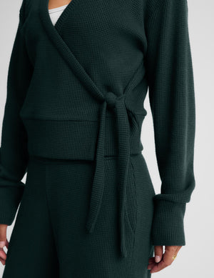 A woman wearing a dark forest green knit wrap cardigan with a tie waist and matching knit pants, photographed against a light gray studio background.