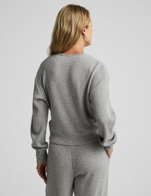 A woman modeling a light gray knit wrap cardigan and matching lounge pants against a plain light gray studio background.