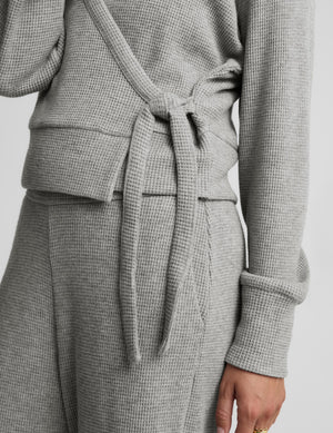 A woman modeling a light gray knit wrap cardigan and matching lounge pants against a plain light gray studio background.