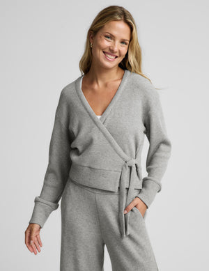 A woman modeling a light gray knit wrap cardigan and matching lounge pants against a plain light gray studio background.