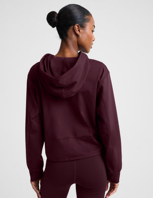 A woman modeling a dark burgundy cropped pullover hoodie with a drawstring hood and a large front kangaroo pocket, paired with matching leggings, against a light gray studio background.