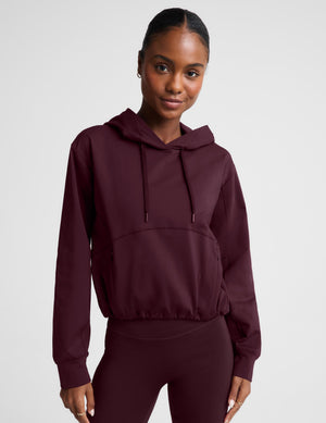 A woman modeling a dark burgundy cropped pullover hoodie with a drawstring hood and a large front kangaroo pocket, paired with matching leggings, against a light gray studio background.