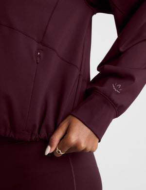 A woman modeling a dark burgundy cropped pullover hoodie with a drawstring hood and a large front kangaroo pocket, paired with matching leggings, against a light gray studio background.