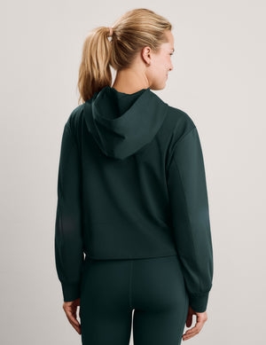 A woman wearing a dark forest green hooded sweatshirt and matching high-waisted leggings, photographed against a neutral light gray studio background.
