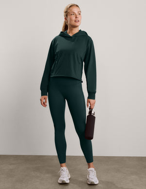A smiling woman wearing a dark forest green hooded sweatshirt and matching high-waisted leggings, photographed against a neutral light gray studio background.
