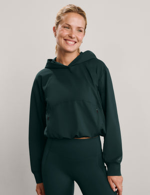 A smiling woman wearing a dark forest green hooded sweatshirt and matching high-waisted leggings, photographed against a neutral light gray studio background.