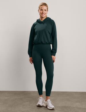 A smiling woman wearing a dark forest green hooded sweatshirt and matching high-waisted leggings, photographed against a neutral light gray studio background.