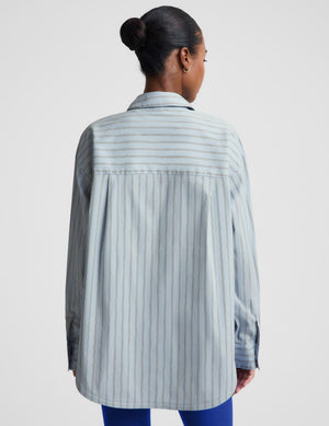 A woman shown from the back wearing an oversized light blue and white vertically striped button-up shirt, paired with blue pants, against a plain light gray studio background.
