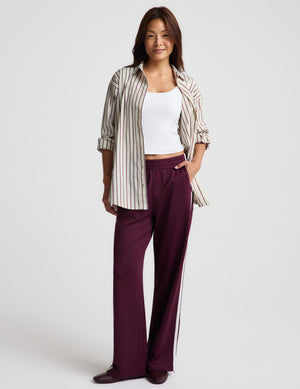 A woman modeling a pair of wide-leg burgundy pants with white side stripes, styled with a white tank top, a striped button-up shirt, and dark loafers against a light gray studio background.