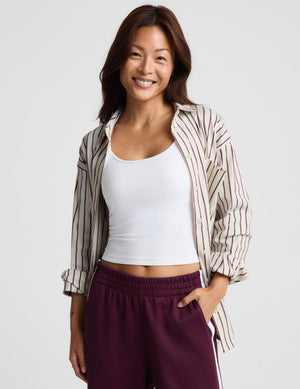 A woman modeling a white ribbed tank top, a striped button-up shirt draped off her shoulders, and burgundy athletic pants with white side stripes, against a light gray studio background.