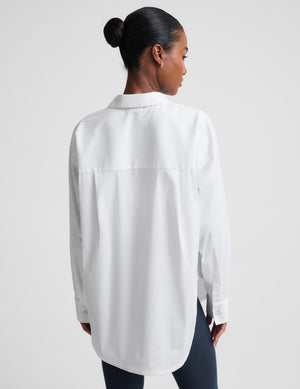 A woman shown from the back wearing an oversized white button-up shirt and dark pants against a light gray studio background.