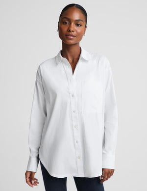 A woman modeling a white long-sleeve button-up shirt with a chest pocket, paired with dark pants, against a light gray studio background.