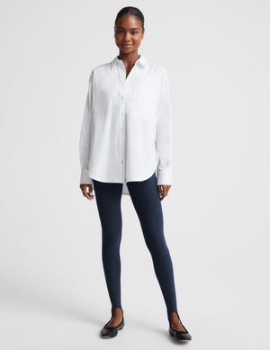 A woman modeling a white long-sleeve button-up shirt paired with dark navy leggings and black flats, standing against a light gray studio background.