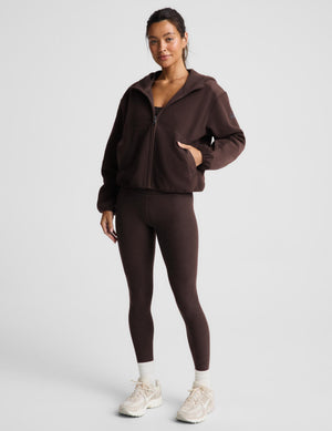 A woman modeling a matching dark brown activewear set consisting of a zip-up hoodie and high-waisted leggings, paired with white socks and beige sneakers, against a light gray studio background.