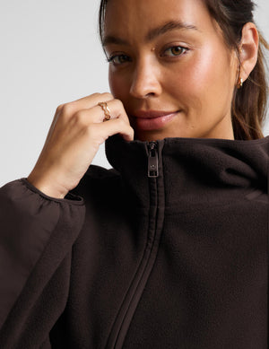 Close-up of a woman wearing a dark brown fleece zip-up hoodie, adjusting the collar with her hand. She is wearing a gold ring and small gold hoop earring against a light gray background.