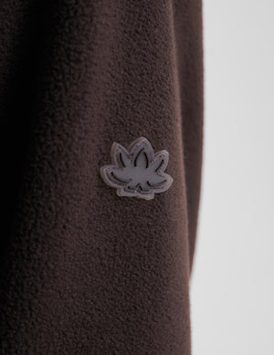 Close-up of a dark brown textured fleece garment featuring a small gray and white embroidered leaf-shaped patch on the chest, set against a light gray background.