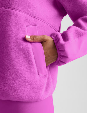 Close-up of a person wearing a bright pink fleece pullover, showing a hand inserted into a front patch pocket against a light gray background.
